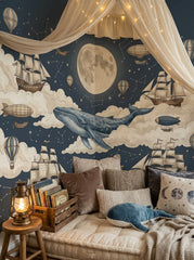 Whimsical whale wall mural with moon, stars, clouds and vintage sailing ships in a cozy children’s reading nook.