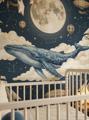 Close-up of blue whale wallpaper with large moon, stars, clouds and vintage hot air balloons behind a white crib.