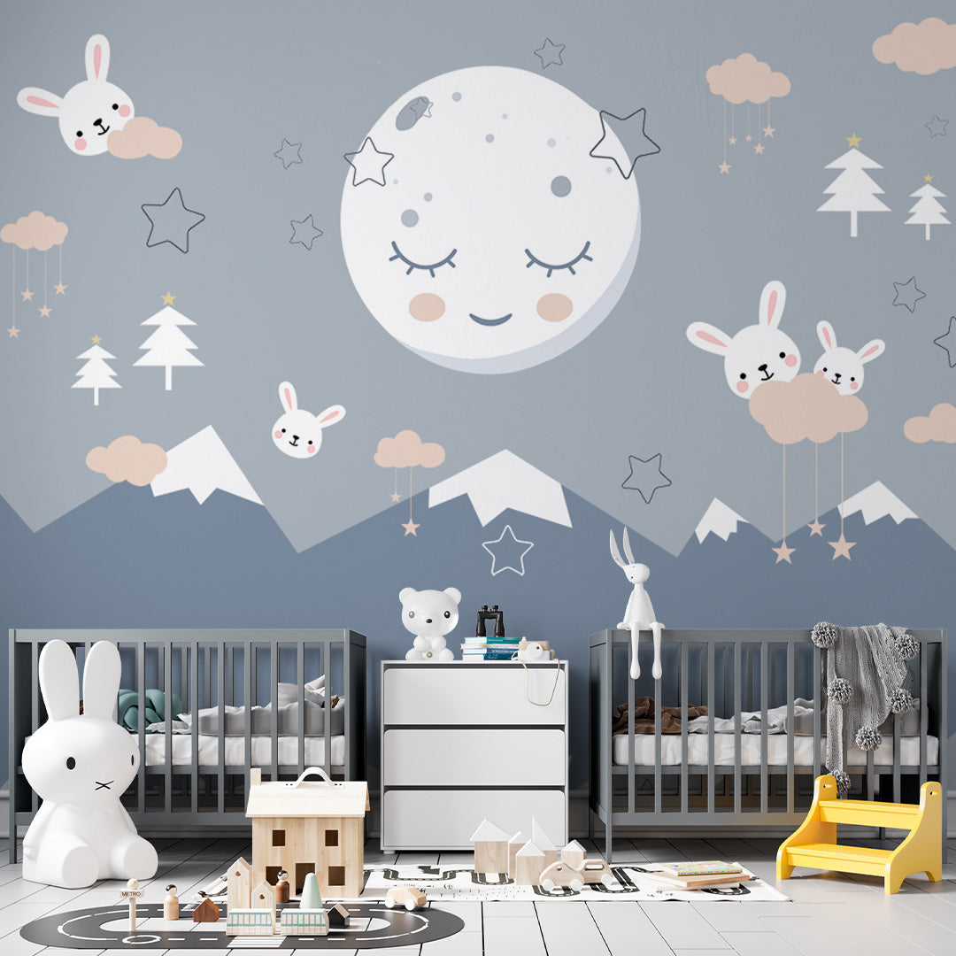 Blue nursery wallpaper with a sleepy moon, little white bunnies, blush clouds, hanging stars, pine trees, and snow-capped mountains.